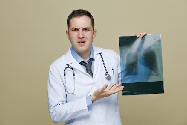 Irritado jovem médico masculino vestindo roupão médico e estetoscópio no pescoço, olhando para a câmera mostrando um tiro de raio-x apontando para ele com a mão isolada no fundo verde-oliva