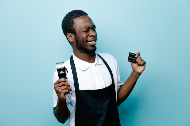 Irritado segurando cartão com máquina de cortar cabelo jovem barbeiro americano africano de uniforme isolado em fundo azul