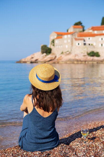 Isla Sveti Stefan, Montenegro 5 de julio de 2021: Mar Adriático. Una niña con un sombrero de paja se sienta en la playa con el telón de fondo de la isla de San Esteban.