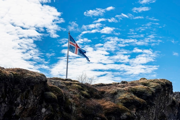 Isländische Flagge weht auf einer felsigen Klippe vor blauem bewölktem Himmel