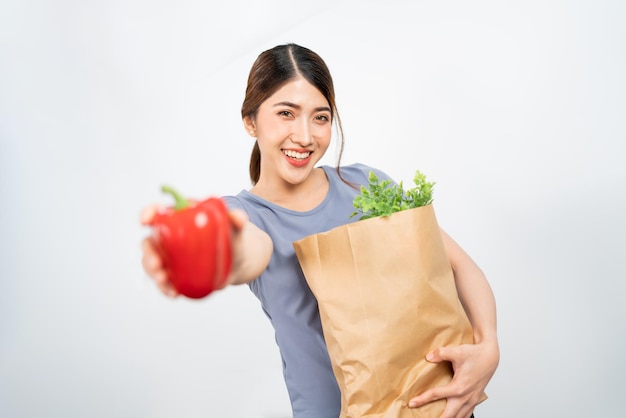 Isoliert ein junges gesundes asiatisches Frauenlächeln und trägt sportliche Kleidung, die Gemüse in einer Papiertüte trägt und der Kamera rote Paprika mit weißem Hintergrund zeigt