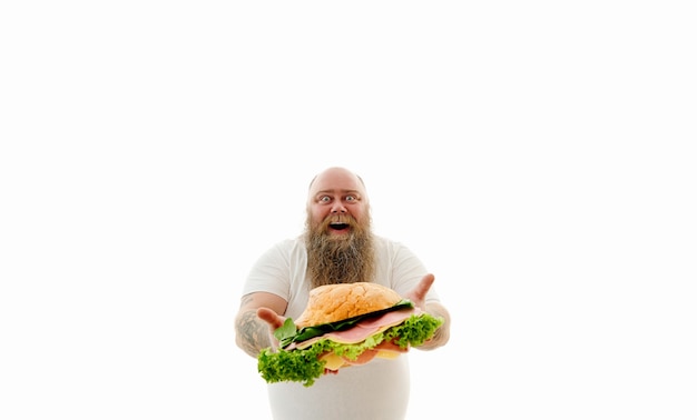 Isoliertes Porträt auf einem weißen Hintergrund. Lustiger übergewichtiger Mann mit tätowierten Armen und langem Bart, der großen Burger mit geschnittenen Würstchen, Käse und Salatblättern zur Kamera zeigt.