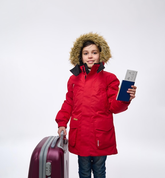 Isoliertes Porträt auf weißem Hintergrund eines hübschen jugendlichen kaukasischen Jungen in leuchtend rotem Parka mit Eco weiterer Kapuze, Blick in die Kamera, die mit einem Koffer posiert und einen Reisepass und eine Bordkarte hält