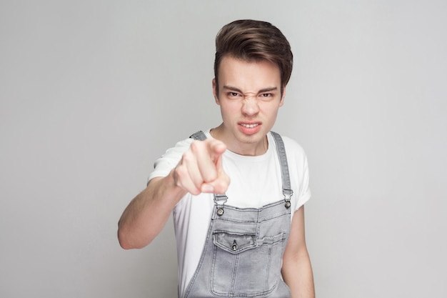 Isso é problema seu. com raiva jovem morena em estilo casual com t-shirt e macacão jeans em pé e olhando para a câmera e acusando. tiro de estúdio interior, isolado em fundo cinza.