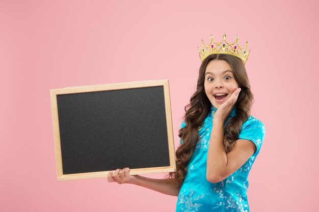 Isso é sensação. Princesa feliz segura o quadro-negro da escola. Garota orgulhosa na coroa de volta à escola. Tabuleiro vazio para promoção ou anúncio, copie o espaço. Criança pequena usa coroa de baile. Celebração do feriado na escola.