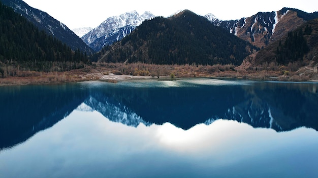 Issyk-Gebirgssee mit Spiegelwasser bei Sonnenuntergang. Wolken, Bäume und Berge werden reflektiert.