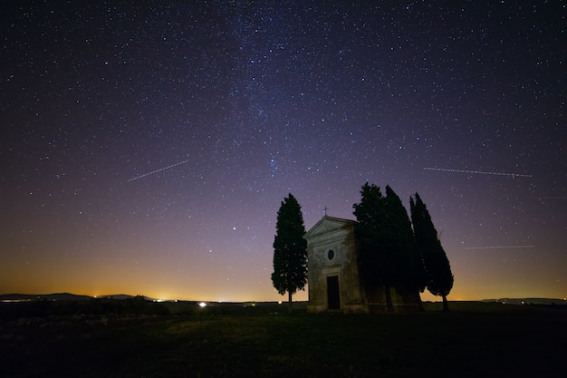 Itália. Toscana. Capela solitária e ciprestes em um campo. Céu noturno com miríades de estrelas