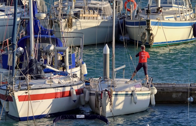 Italien, Sizilien, Mittelmeer, Marina di Ragusa (Provinz Ragusa); 8. Februar 2021, Frau macht Gymnastik und Segelboote im Hafen – EDITORIAL