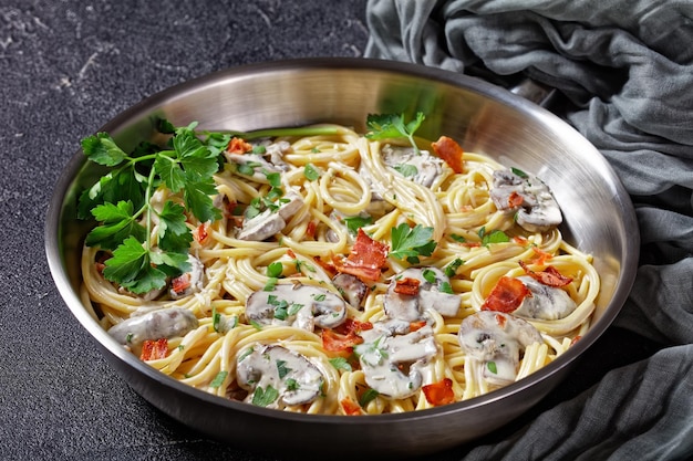 Italienische Pasta mit Cremini-Pilzsauce aus Sahne und Parmesankäse mit knusprigem Speck und frischer Petersilie, serviert auf einer Metallpfanne auf dunklem Betonhintergrund, Draufsicht, Nahaufnahme