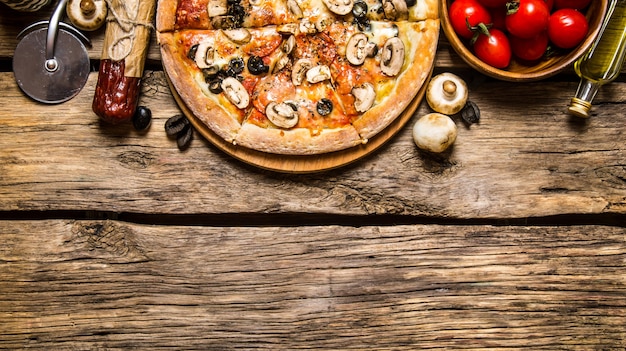 Italienische Pizza und verschiedene Zutaten - Fleisch, Pilze, Tomaten und Olivenöl. Auf hölzernem Hintergrund.