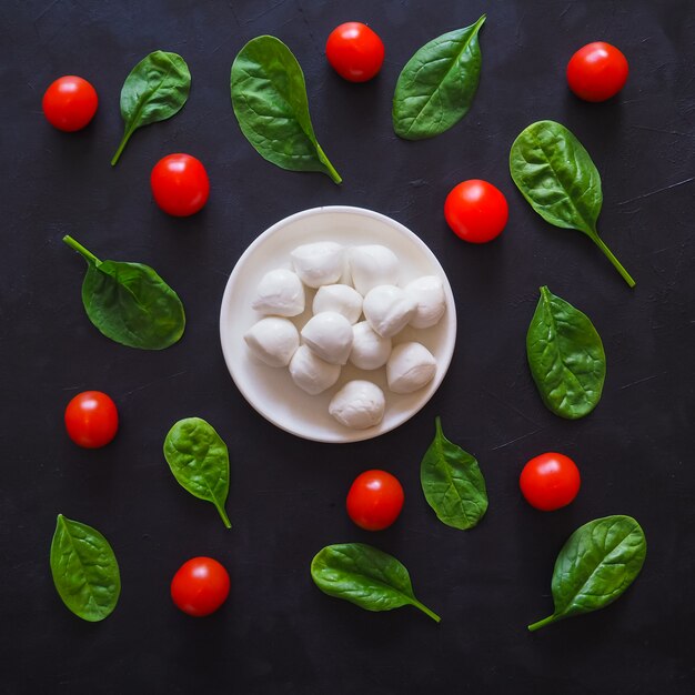 Italienischer Mozzarella-Käse, Kirschtomaten und Spinatblätter auf einem schwarzen Tisch.