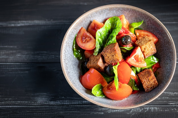 Italienischer Salat aus geschnittenen Tomaten, Basilikumblättern, Oliven, großen Croutonstücken. In einer tiefen Platte auf einem dunklen Hintergrund.