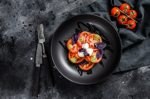 Italienischer traditioneller Caprese-Salat. Kirschtomaten, Mozzarella, Basilikum, Pesto-Sauce. schwarzer Hintergrund. Draufsicht