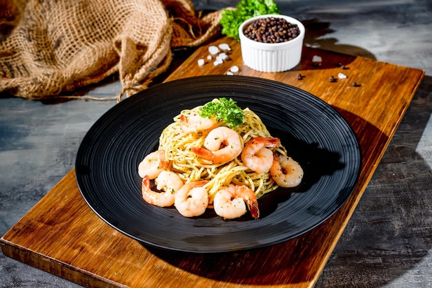 Italienisches Essen Garnelen Aglio Olio Nudeln mit schwarzem Pfeffer in einem Gericht auf Holzbrett Seitenansicht auf dunklem Hintergrund