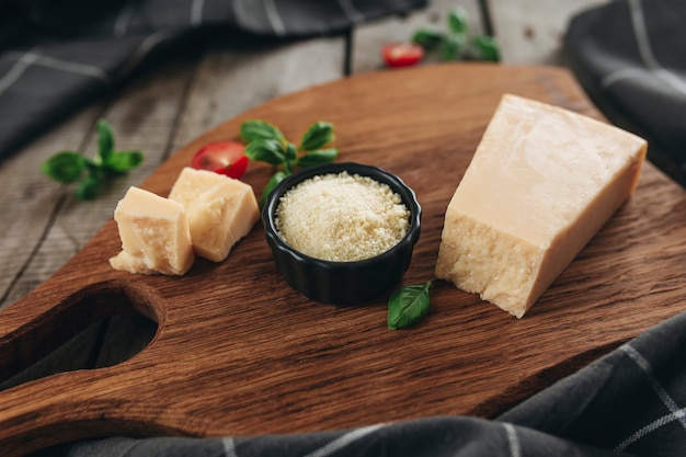 Italienisches Küchenkonzept. Schneidebrett, Parmesan-Käse-Stücke, geriebener Käse in einer kleinen schwarzen Schüssel, Kirschtomaten, Basilikumblätter, Küchentuch auf Holztisch. Gesundes Esskonzept