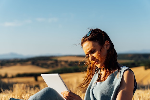 J-Frau mittleren Alters, die von der Tablette mit Sonnenbrille im Haar liest, die in einem Getreidefeld sitzt. Arbeiten. lesen.