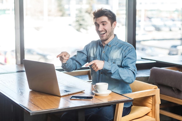 Ja, du bist es! Seitenansichtporträt eines glücklichen, gutaussehenden, bärtigen jungen Freiberuflers im blauen Jeanshemd sitzt im Café und führt einen Videoanruf auf dem Laptop durch, zeigt mit dem Finger und wählt den Begleiter aus. drinnen