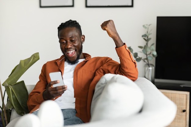 Ja, tolle Neuigkeiten Überglücklicher afroamerikanischer Mann, der ein Smartphone benutzt und die geballte Faust schüttelt, sitzt auf dem Sofa