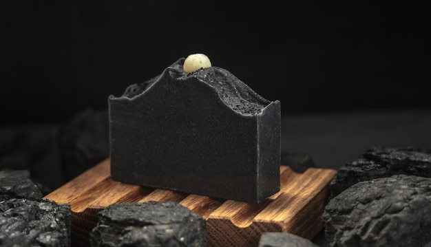 Jabón de carbón negro natural en una jabonera de madera rodeada de carbones Concepto de hacer y usar jabón orgánico y cosméticos regalos hechos a mano ecológicos