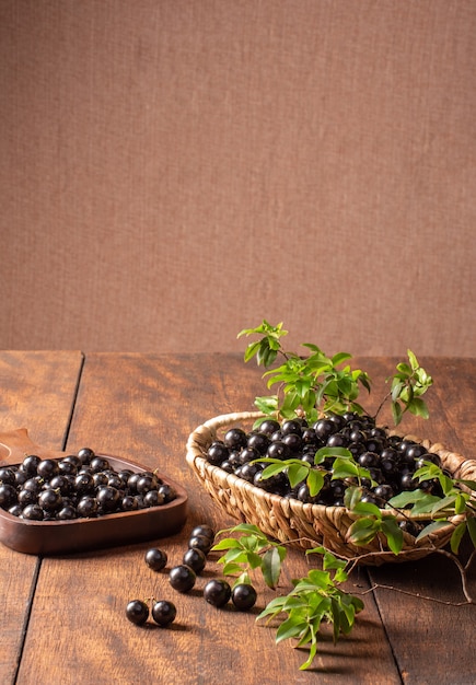 Jabuticaba, Jabuticabas recién cosechadas y aún no lavadas, colocadas en una canasta de paja y cuenco de madera sobre madera rústica, enfoque selectivo.