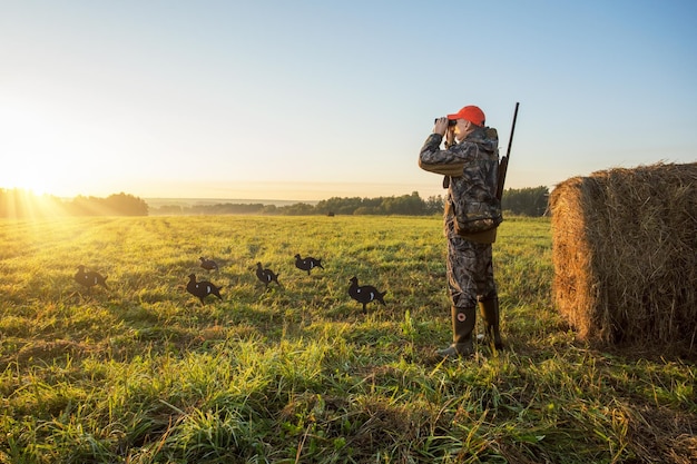 Jäger in Tarnung mit Gewehr jagt im Morgengrauen Birkhühner. Jäger schaut durch ein Fernglas auf das Herbstfeld