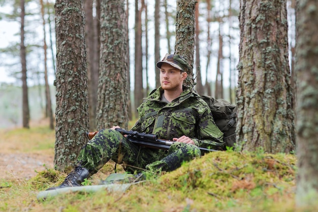 Jagd-, Kriegs-, Armee- und Menschenkonzept - junger Soldat, Ranger oder Jäger mit Waffe und Rucksack, der im Wald ruht