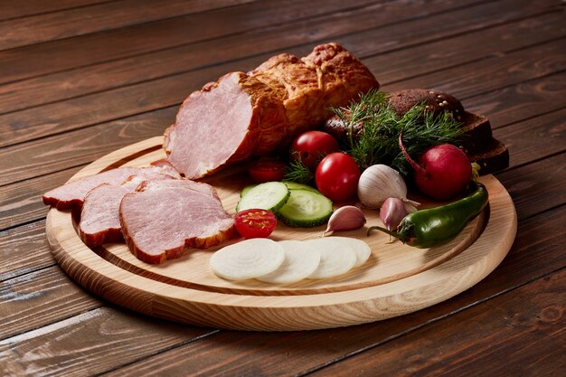 Foto jamón aderezado con verduras, frutas y hierbas con salsas y pan en una placa de madera sobre una mesa de madera oscura.