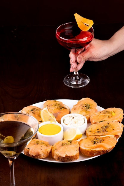 Jantar com prato de bruschetta de salmão defumado com pepino e queijo mussarela mão feminina pegando copo de coquetel no fundo desfocado