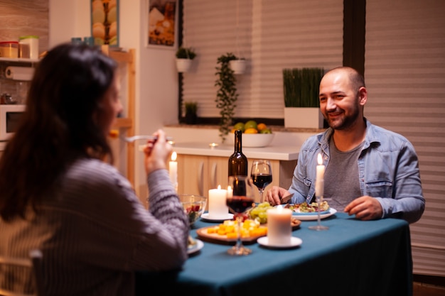 Jantar romântico com o homem em primeiro plano, sentindo-se feliz com a esposa na sala de jantar. Relaxe as pessoas felizes tilintando, sentadas à mesa na cozinha, apreciando a refeição, comemorando o aniversário.