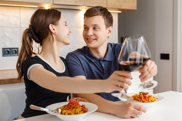 Jantar romântico de dia dos namorados em casa. Casal feliz sorrindo, tilintando copos e comendo macarrão com vinho na cozinha. Conceito de estilo de vida doméstico, amor, casamento feliz e união.