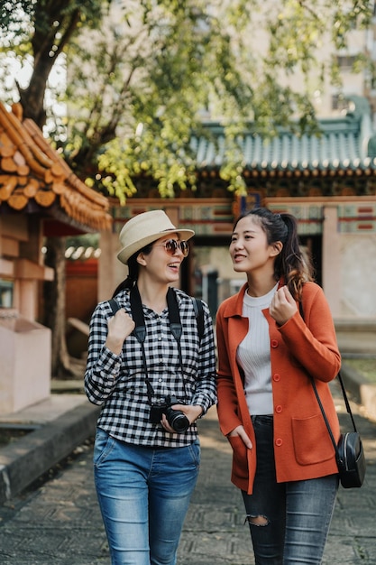 japanische reisende entspannen sich beim gehen an einem sonnigen tag im garten im chinesischen stil im konfuzius-tempel in peking china. Junge Mädchen reden, plaudern, lachen im Park mit riesigem Geschichtstor im Rücken von Baum