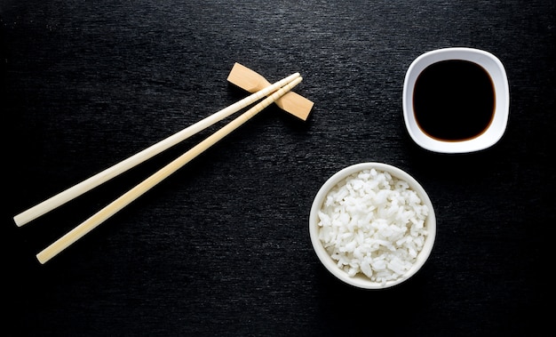 Japanische Sushiessstäbchen über Sojasoßenschüssel, Reis auf schwarzem Hintergrund