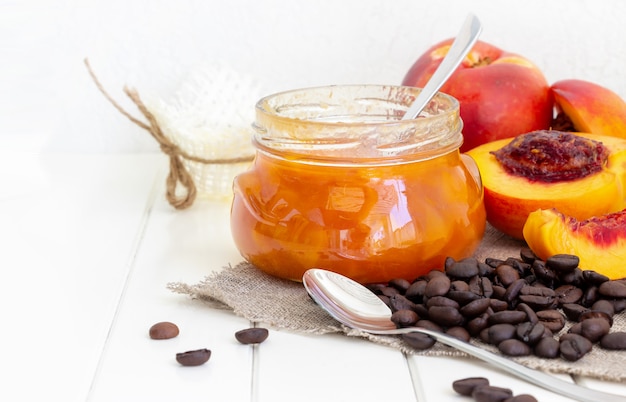 Jar com geléia de pêssego ou nectarina. Frutos de meia nectarina. Grãos de café no guardanapo de serapilheira. Espaço de madeira branco. Foco seletivo. Copie o espaço.