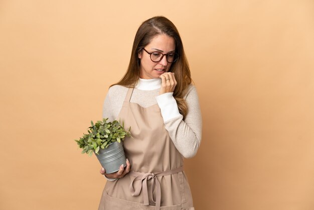 Jardineira de meia-idade segurando uma planta isolada em bege, tendo dúvidas