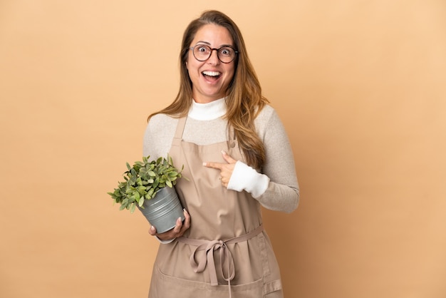 Jardineira de meia-idade segurando uma planta isolada em uma parede bege com expressão facial surpresa