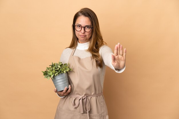 Jardineira de meia-idade segurando uma planta isolada em uma parede bege fazendo gesto de pare