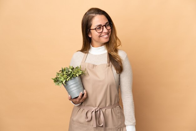 Jardineira de meia-idade segurando uma planta isolada em uma parede bege, olhando para o lado e sorrindo