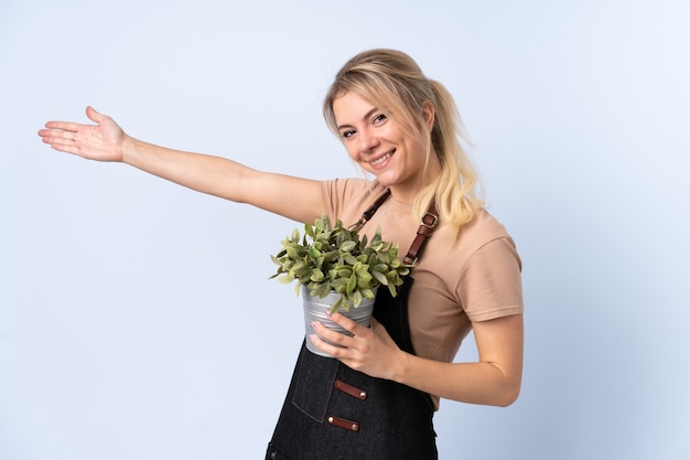 Foto jardineira loira segurando uma planta sobre uma parede isolada e estendendo as mãos para o lado para convidá-la a vir