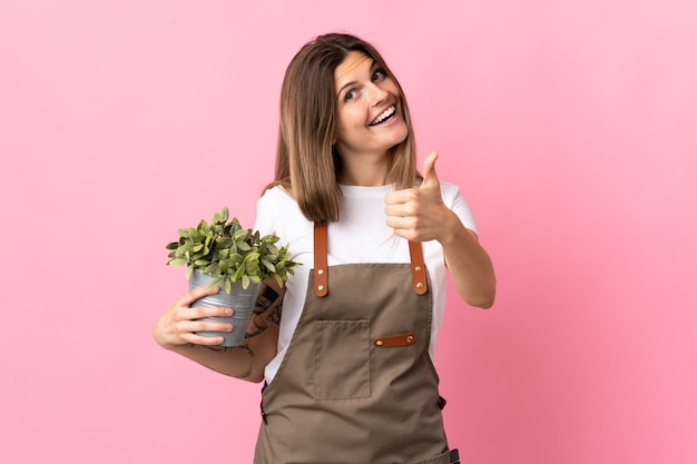 Jardineira segurando uma planta isolada em um fundo rosa com o polegar para cima porque algo bom aconteceu