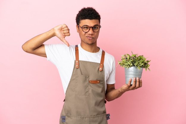 Jardineiro africano segurando uma planta isolada em um fundo rosa, mostrando o polegar para baixo com expressão negativa