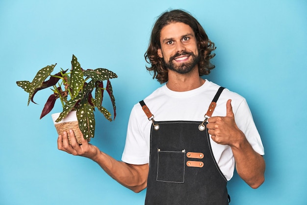 Jardineiro de cabelo longo com barba segurando uma planta sorrindo e levantando o polegar para cima