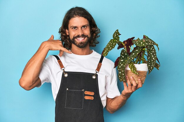 Foto jardineiro de cabelos longos com barba segurando uma planta mostrando um gesto de chamada de telefone móvel com os dedos