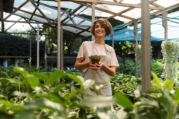 Jardineiro de mulher bonita e fofa em pé sobre plantas de flores em uma estufa segurando plantas