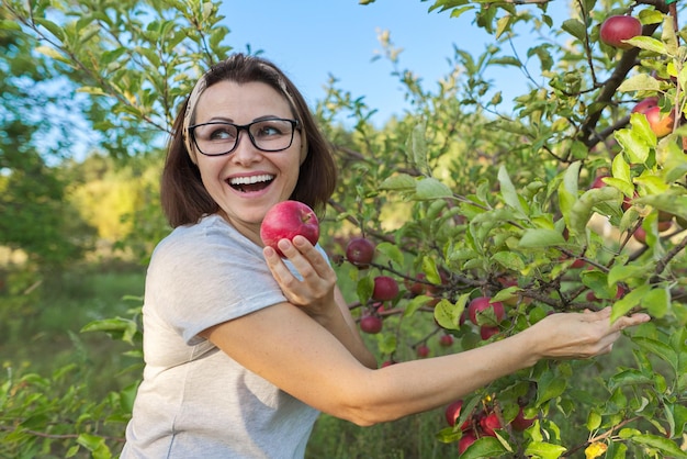 Jardineiro de mulher com maçã vermelha recém-colhida à disposição, o plano de fundo é árvore com maçãs. A fêmea come maçã natural e ecologicamente correta, cultivada na horta, copie o espaço