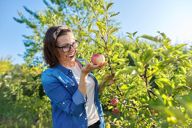 Jardineiro feminino sorridente perto de macieira com maçãs vermelhas maduras, segura a maçã na mão, copie o espaço. hobbies e lazer, horta doméstica cultivando alimentos orgânicos naturais saudáveis, conceito de jardinagem