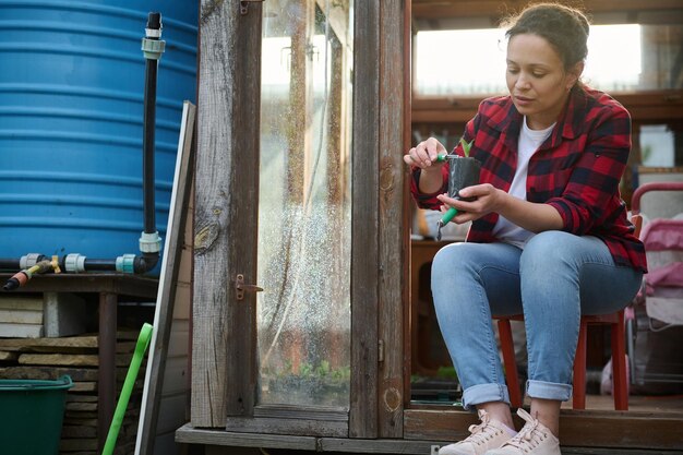 Una jardinera bonita que usa un rastrillo de jardinería y una pala afloja y cava el suelo negro con un brote sentado en la entrada de su viejo invernadero de madera