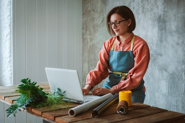 Foto una jardinera con gafas y un delantal, sentado en una mesa de madera contra una losa de hormigón, usando una computadora portátil para trabajar