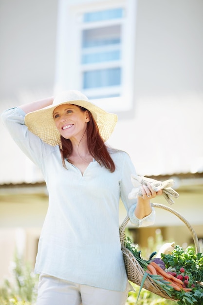 La jardinería es mi forma de relajarme Una hermosa mujer sostiene sus guantes de jardinería y una canasta de verduras recién cortadas mientras está de pie en su jardín