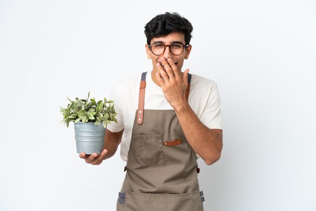 Jardinero argentino sosteniendo una planta aislada de fondo blanco feliz y sonriente cubriendo la boca con la mano