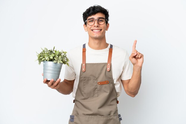 Jardinero argentino sosteniendo una planta aislada de fondo blanco mostrando y levantando un dedo en señal de lo mejor